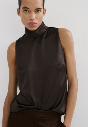 Person iført en ærmeløs mørkebrun højhalset top og matchende bukser, stående mod en enkel lys baggrund.