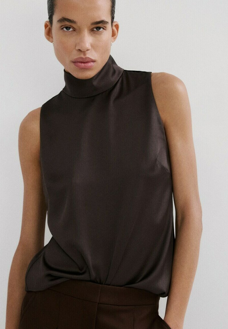 Osoba ubrana w ciemnobrązowy, bezrękawnik z golfem oraz dopasowane spodnie, stojąca na jasnym, jednolitym tle.