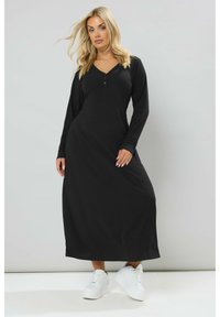 Abito maxi nero a maniche lunghe con texture a coste, scollo a V e dettagli con bottoni, abbinato a sneakers bianche su uno sfondo chiaro.