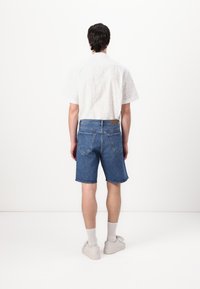 Calções de ganga em azul médio com uma bainha desgastada, combinados com uma camisa branca de mangas curtas com um padrão subtil, e meias cinzas com sapatos brancos.