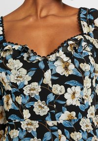 Robe noire à motifs floraux avec un col volanté, des motifs floraux bleus et blancs, et des bordures noires. Le matériau semble léger et texturé.