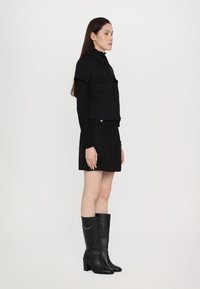 Veste noire cropped avec poches poitrine doubles, associée à une mini-jupe noire texturée et des bottes noires en cuir à talons montant jusqu'aux genoux.
