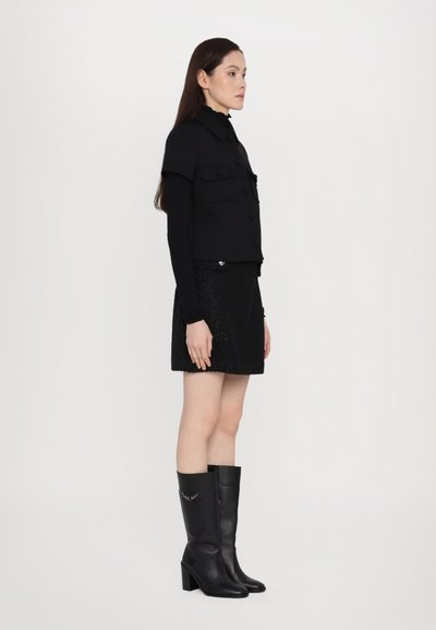 Black cropped jacket with dual chest pockets, paired with a textured black mini skirt and knee-high, heeled black leather boots.