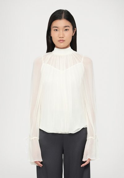 Femme portant un chemisier blanc transparent à manches longues avec des poignets volantés et un col montant, associé à un pantalon droit foncé, sur un fond uni.