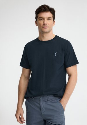 Mand står med den ene hånd i lommen iført en mørkeblå kortærmet t-shirt med lille golfspillerlogo og grå bukser.