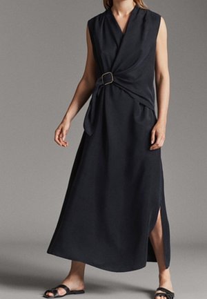 Robe portefeuille noire sans manches avec une fente latérale, maintenue par une boucle rectangulaire, portée avec des sandales plates noires à bout ouvert sur un fond uni.