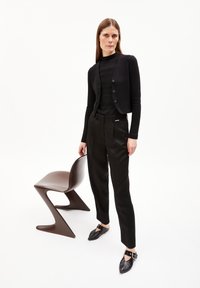 Black cropped cardigan with buttons, fitted black top, tailored black trousers, and pointed black shoes. Brown modern chair beside model.