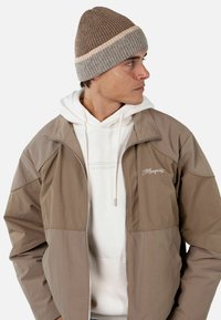Chaqueta marrón con cremallera y un diseño en bloques de color marrón claro y beige, llevada sobre una sudadera blanca con capucha. Gorro marrón acanalado con una franja beige.