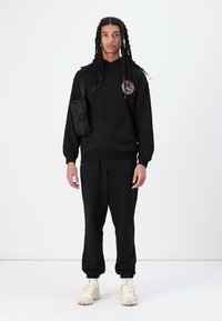 Svart hettegenser med logo på brystet, kombinert med matchende joggebukser. Tilbehør inkluderer en liten svart crossover-veske og lyse sneakers.