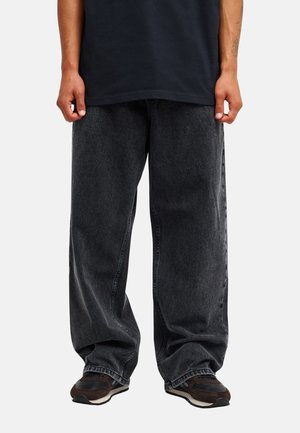 Personne portant un jean noir ample délavé et des baskets noires, debout devant un fond blanc uni.