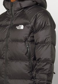 Veste matelassée noire avec capuche, fermeture éclair avant, poche zippée sur la poitrine et logo The North Face sur la partie gauche de la poitrine.