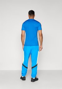 Camiseta atlética azul de manga corta y pantalones jogger azules con paneles laterales de malla amarilla y rayas negras. Zapatillas deportivas negras completan el conjunto.