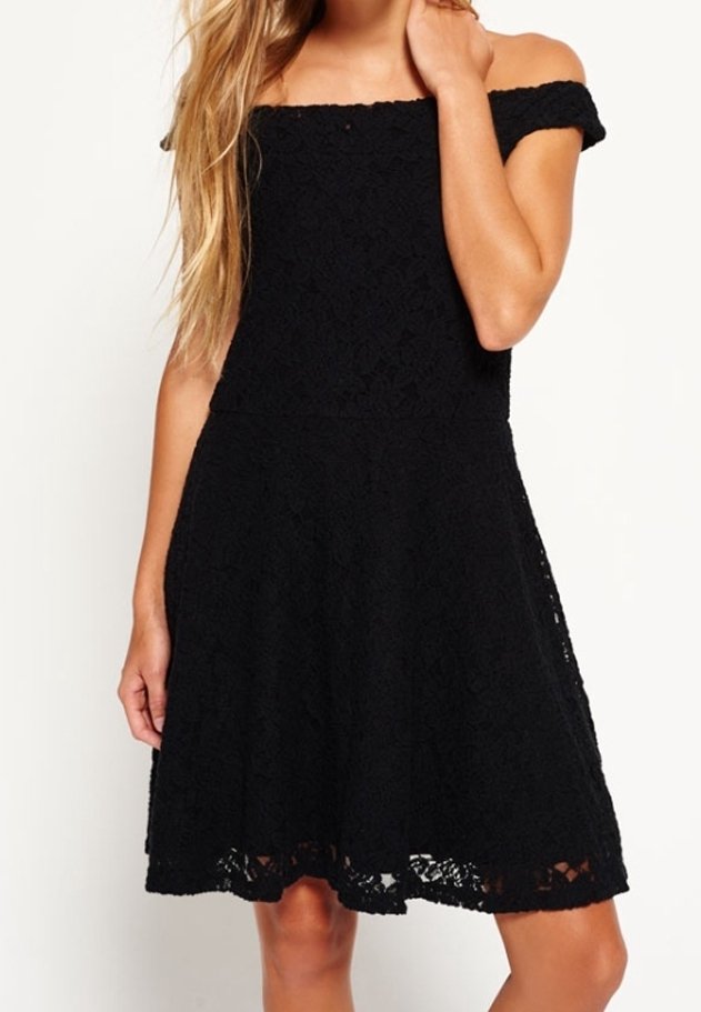 Femme portant une robe noire en dentelle épaules dénudées avec un corsage ajusté et une jupe évasée, debout devant un fond uni.