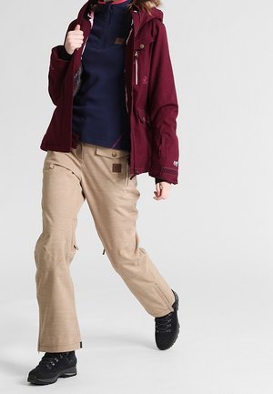 Giacca imbottita color amaranto con cappuccio, maglione blu scuro e pantaloni da sci beige. Stivali da trekking neri completano l'abbigliamento da esterno.