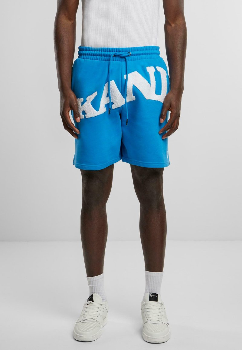 Tirkīza krāsas sporta šorti ar baltu pūkainu "KAND" logo, ar aukliņu jostas daļu un sānu kabatām, izgatavoti no mīksta, tekstūrēta auduma.