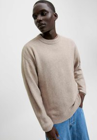 Beige kofta i mjukt tyg med ribbad rund hals och nederkant; avslappnad passform och långa ärmar; kombinerad med blå denimshorts.