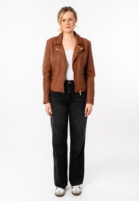 Giacca da moto in pelle marrone con cerniera asimmetrica, indossata sopra una maglietta bianca, abbinata a jeans neri a gamba larga e sneaker bianche.