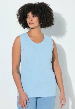 Vrouw met krullend haar, gekleed in een mouwloos lichtblauw topje en een blauwe broek, staand en zijwaarts kijkend tegen een witte achtergrond.