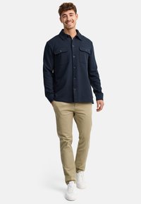Chemise boutonnée bleu marine avec deux poches poitrine, associée à un pantalon beige et des baskets blanches. La chemise a un col et un tissu texturé.