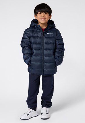 Bambino sorridente con giacca puffer blu navy con cappuccio, pantaloni blu navy e sneakers bianche, in piedi con le mani nelle tasche della giacca.