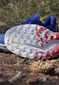 Blue athletic shoe with a textured white and red outsole featuring a geometric pattern and a yellow logo. Surrounded by natural ground debris.