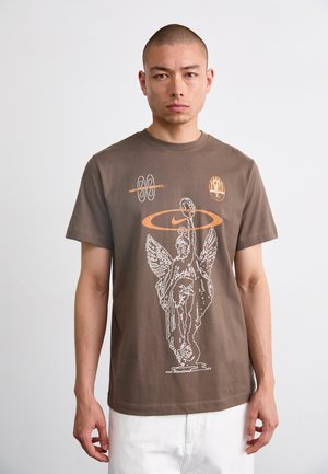 Ruda medvilnė marškinėliai su balta grafika, vaizduojančia skulptūrą su sparnais ir oranžiniais akcentais, įskaitant Nike logotipą ir tekstą.