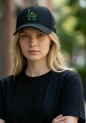 Jeune femme aux longs cheveux blonds portant un t-shirt noir et une casquette noire avec un logo floral LA, debout en extérieur avec un fond vert flou.