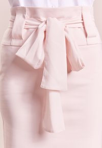 Jasnoróżowa spódnica z szerokim pasem w talii i związana kokardką z przodu. Tkanina jest gładka, o przemyślanej konstrukcji i subtelnej fakturze.