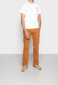 White cotton t-shirt with a small red logo, paired with orange cargo pants featuring side pockets, and beige sneakers with black accents.