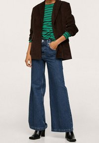 Žena v širokých modrých džínách, černých kožených botách na podpatku, dlouhém tričku s green a černými pruhy a tmavě hnědém kordurovém blejzru.