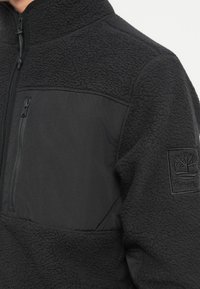 Svart fleece-pullover med glidelås på brystlommen, strukturert kropp, glatte paneler og Timberland-logo på ermet.