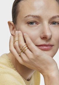 Gold rings in various designs, featuring textured and smooth accents, worn on fingers touching a face against a neutral background.