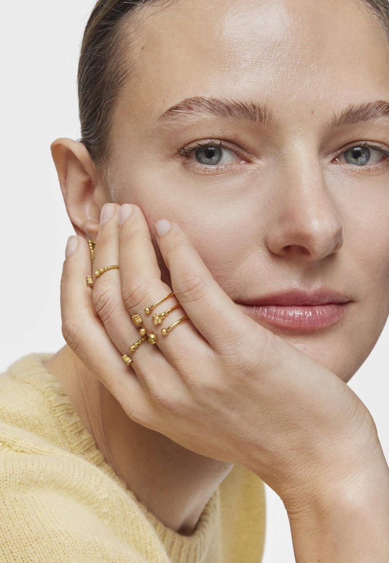 Gold rings in various designs, featuring textured and smooth accents, worn on fingers touching a face against a neutral background.