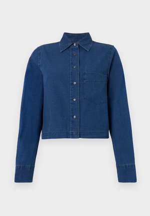 Henkilö, jolla on päällään lyhyt tummansininen napillinen paita, beige polveen ulottuvat shortsit, mustat teräväkärkiset tasapohjaiset kengät ja kantamassa ruskeaa olkalaukkua.