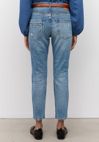 Jeans de mezclilla azul con un corte ajustado, dobladillo deshilachado y dos bolsillos traseros. Combinados con un cinturón de cuero marrón y zapatos negros de slip-on.