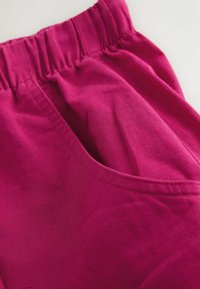 Pantaloni in tessuto rosa acceso con elastico in vita e cuciture curve sulla tasca laterale in primo piano.