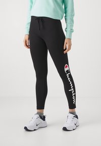Czarne legginsy o gładkiej fakturze, z białym logiem "Champion" i kontrastującym czerwonym akcentem na lewym nogawce. Noszone z jasnoniebieską bluzą.