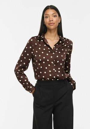 Femme aux longs cheveux foncés portant un chemisier marron à boutons avec des pois blancs et un pantalon noir, debout les mains dans les poches.