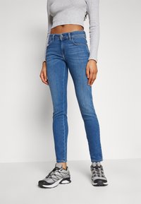 Jeans skinny de mezclilla azul claro con cintura alta, que cuentan con cinco bolsillos y costuras sutiles. Combinados con zapatillas deportivas grises.