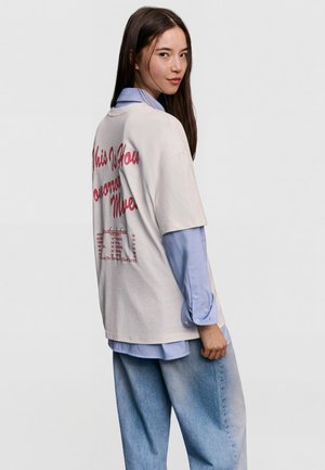 Mladá žena v volném bílém tričku s červeným nápisem přes světle modrou košili na knoflíky a volné modré džíny, stojící na jednobarevném pozadí.