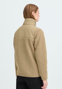 Beige fleecejakke med en tekstureret overflade og en glat, cremefarvet øverste sektion. Har høj krave og lange ærmer.