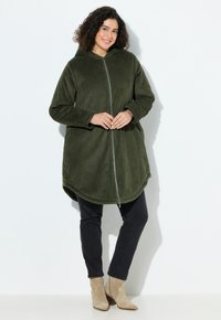 Olijfgroen corduroy zip-up jas met capuchon, met lange mouwen en een asymmetrische zoom, gecombineerd met zwarte jeans en beige enkellaarzen.