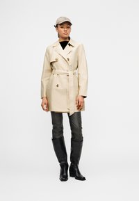 Γυναίκα που φοράει μπεζ παλτό με ζώνη, γκρι καπέλο, ξεθωριασμένο γκρι τζιν και μαύρες μπότες με τακούνι μέχρι το γόνατο, στέκεται μπροστά σε λευκό φόντο.