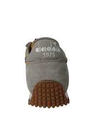 Sneaker grigia in suede con una suola in gomma marrone texturizzata. Presenta un logo ricamato bianco e l'anno "1975" sul tallone. Design curvo.