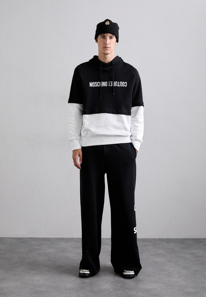 Felpa nera corta con scritte bianche, indossata su una maglietta bianca a maniche lunghe, abbinata a pantaloni sportivi neri larghi e un berretto nero.