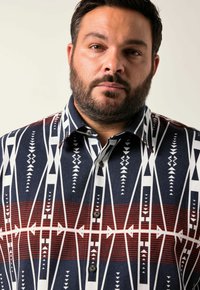 Hombre con barba que lleva una camisa de botones con un patrón geométrico azul, blanco y rojo, contra un fondo claro y liso.