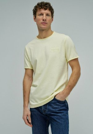 Hombre con cabello rizado, vistiendo una camiseta amarilla pálida de "Salsa Jeans" y jeans azules, de pie con una mano en el bolsillo frente a un fondo liso.