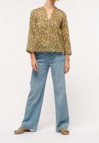 Blouse légère à motif floral en moutarde et vert, associée à un jean denim clair à jambes larges et des sandales plates vert olive.