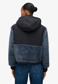 Giacca con cappuccio caratterizzata da una combinazione di pelliccia blu scuro testurizzata e tessuto nero liscio, con polsini a costine e design corto.