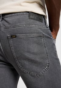 Gray denim jeans with a classic five-pocket design. Features a black leather patch at the waistband and yellow logo tag on the back pocket.